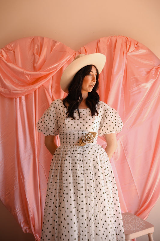Woman in a polka dot dress and hat standing in front of pink draped fabric