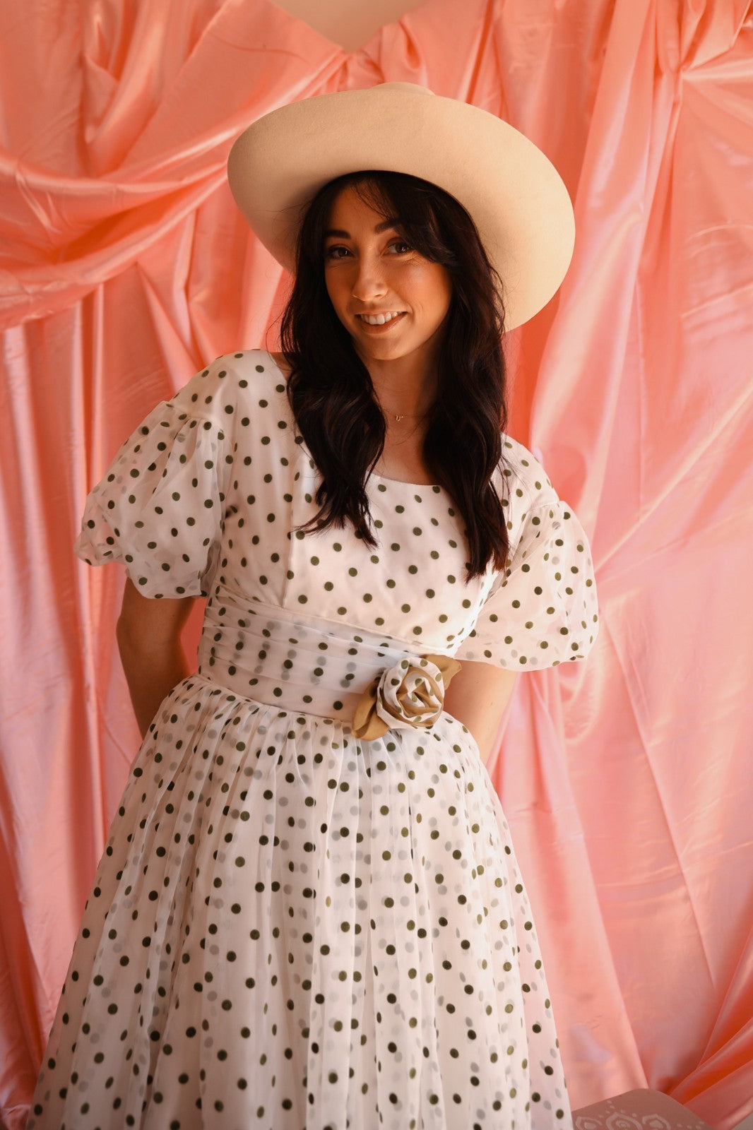 Woman wearing a polka dot dress and wide-brimmed hat against a pink curtain background
