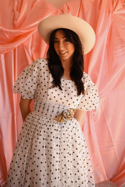 Woman wearing a polka dot dress and wide-brimmed hat against a pink curtain background