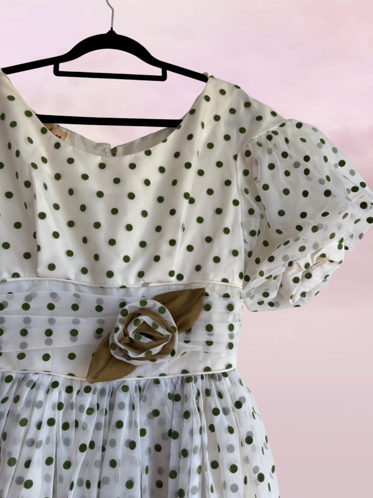 White dress with green polka dots and a brown belt on a hanger against a pink background