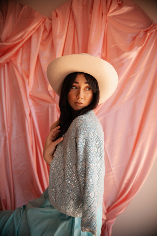 Woman wearing a wide-brimmed hat and light blue patterned top against a pink draped curtain.