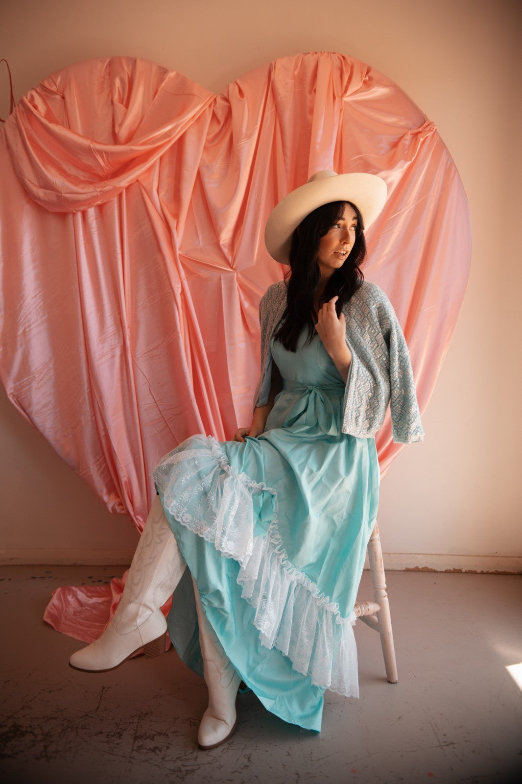 Woman in a light blue dress and white hat sitting on a stool against a pink curtain backdrop.