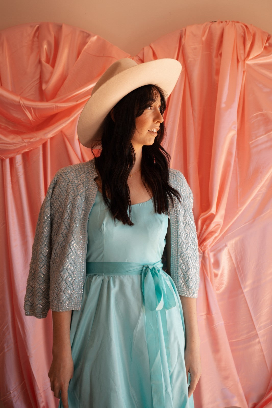 Woman wearing a light blue dress and beige hat against a pink draped backdrop