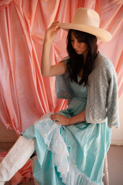Woman wearing a light blue dress with lace details and a gray cardigan, holding a beige hat against a pink curtain background.