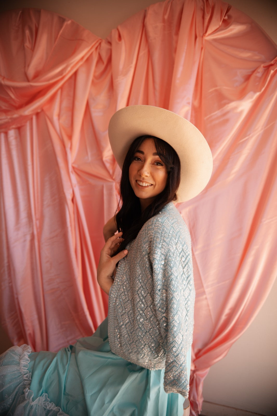 Woman wearing a white hat and light blue dress against a pink draped backdrop