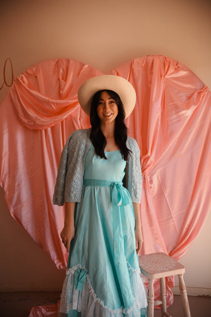 Woman in a light blue dress and beige hat standing in front of a pink curtain