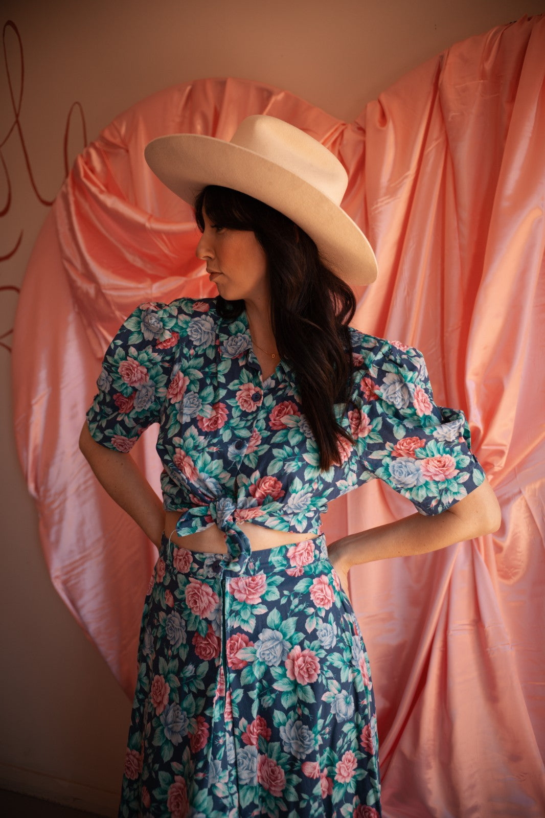 Woman wearing a floral dress and wide-brimmed hat against a pink curtain backdrop