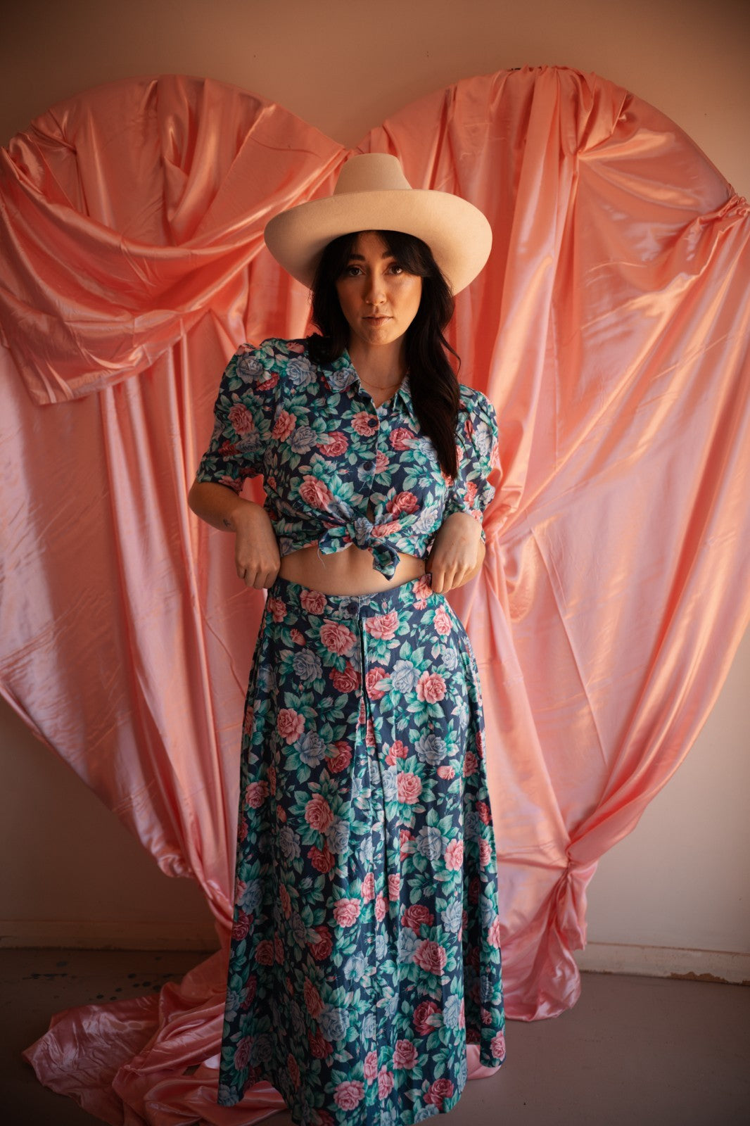 Woman in a floral dress and hat standing against a pink draped curtain backdrop