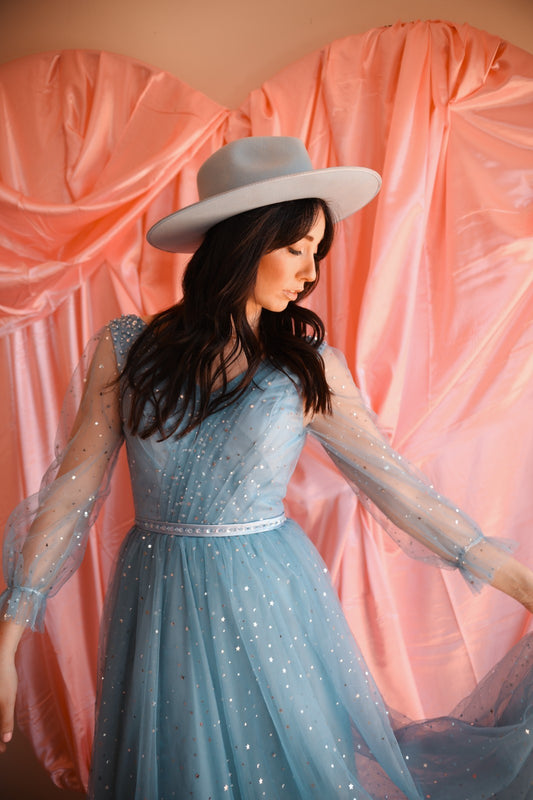 Woman in a blue dress with sheer sleeves and a white hat against a pink curtain backdrop