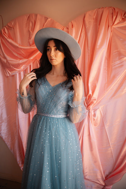 Woman in a blue dress and gray hat standing against a pink curtain backdrop