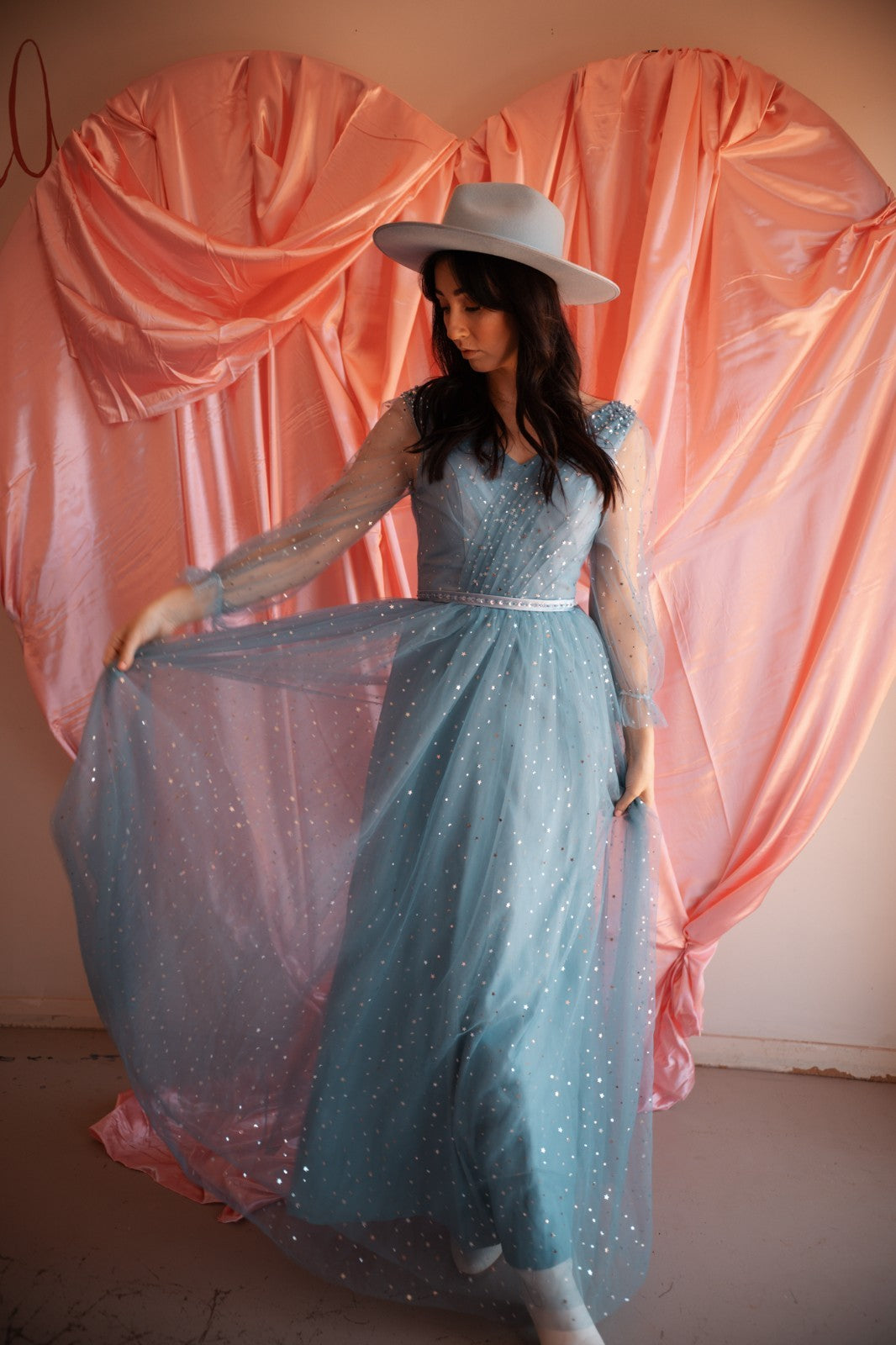 Woman in a long, light blue dress with a white hat standing against a pink curtain backdrop.