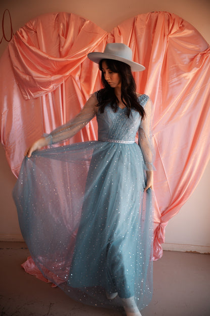 Woman in a long, light blue dress with a white hat standing against a pink curtain backdrop.
