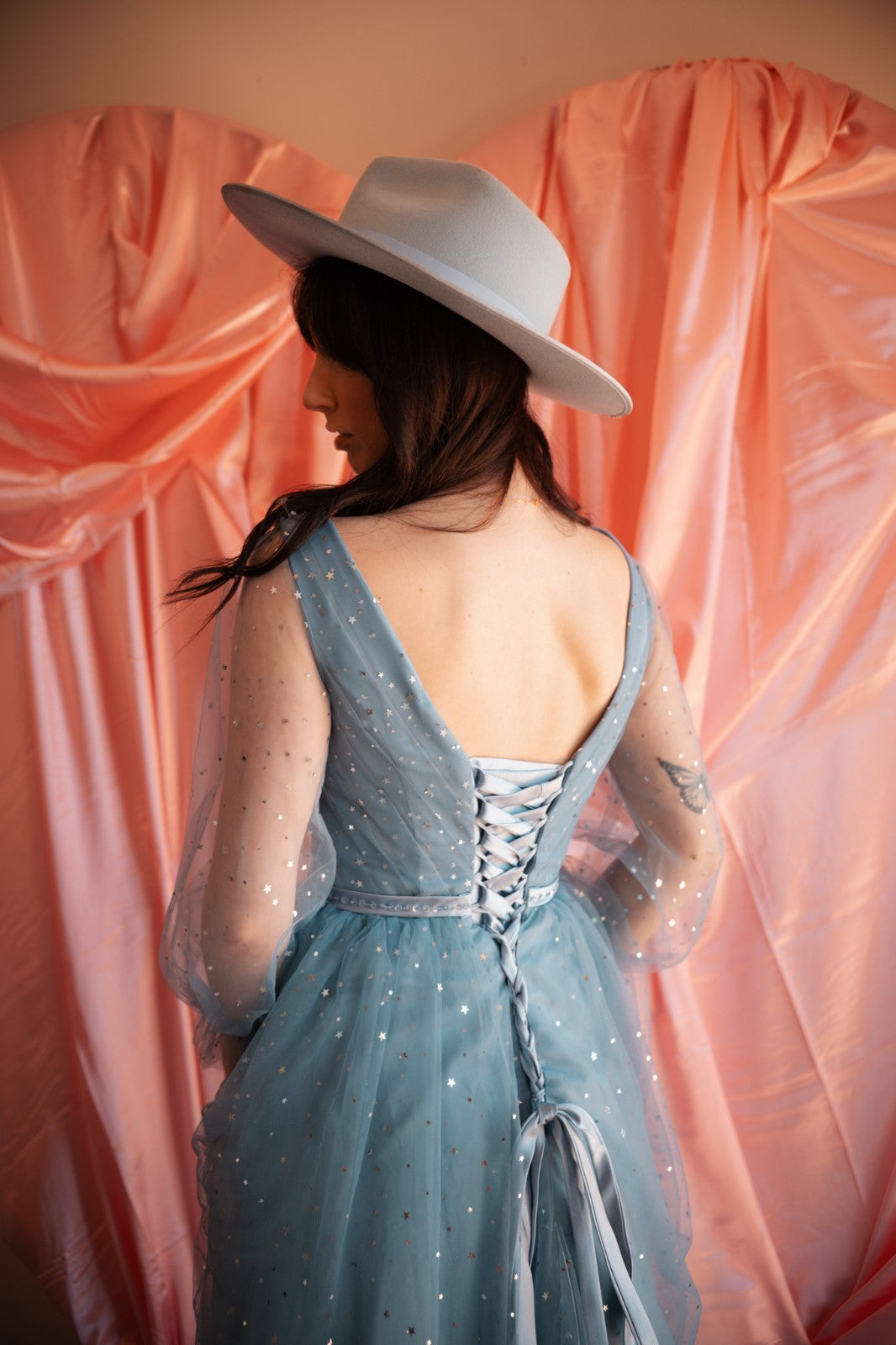 Woman wearing a light blue dress with sheer sleeves and a white hat against a pink curtain backdrop