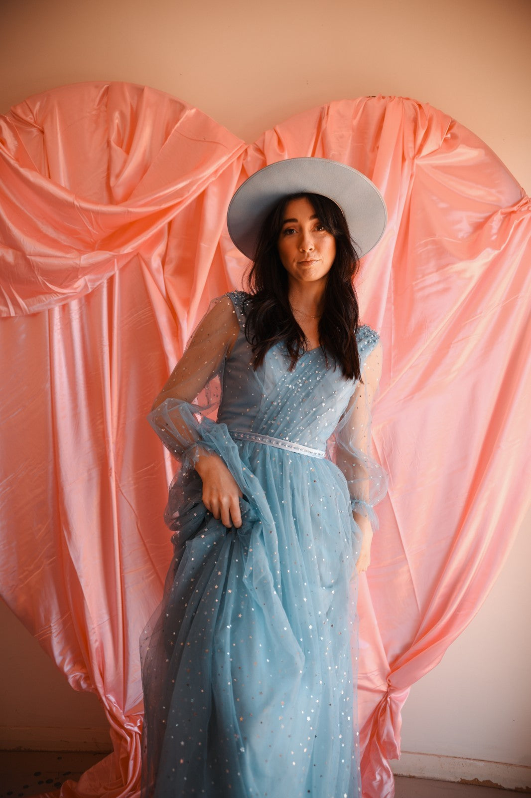 Woman in a blue dress and hat standing in front of pink fabric