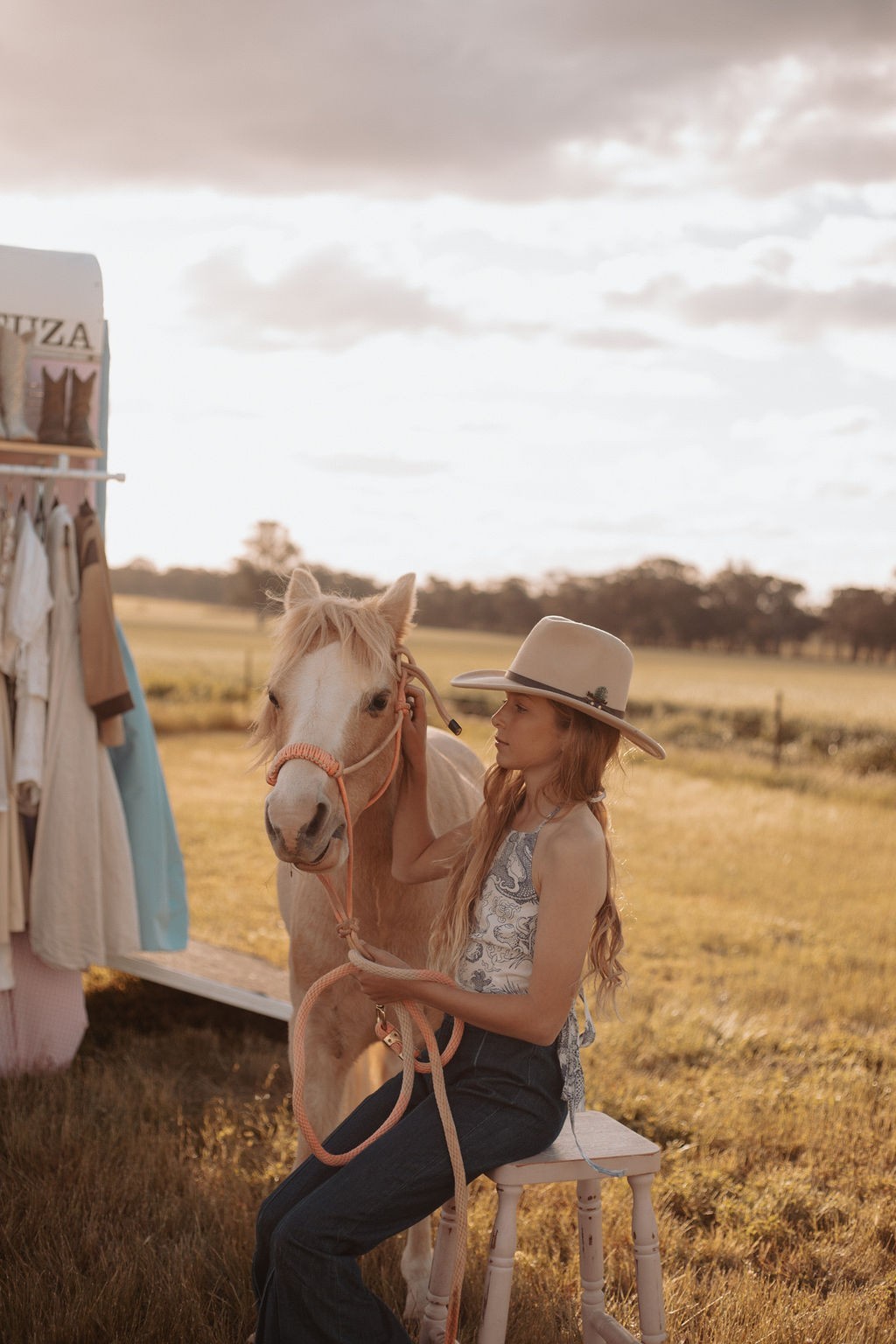 Woman sitting on a stool next to a horse in a field with a trailer in the background.