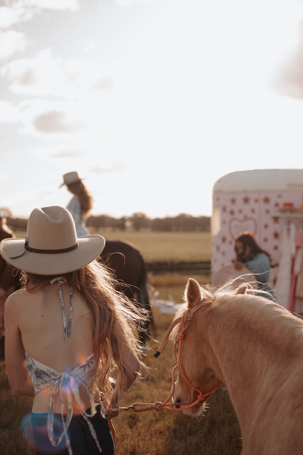 Person in a cowboy hat standing next to a horse with a field and people in the background.