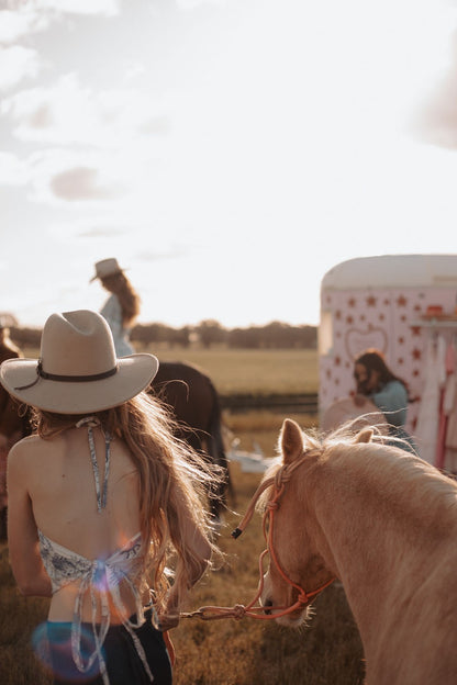 Person in a cowboy hat standing next to a horse with a field and people in the background.
