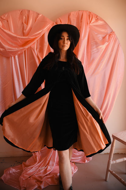 Woman in a black dress with a long pink train standing against a pink curtain backdrop.