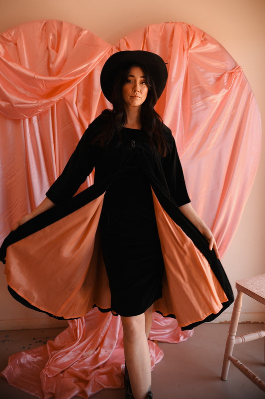 Woman in a black dress with a long pink train standing against a pink curtain backdrop.