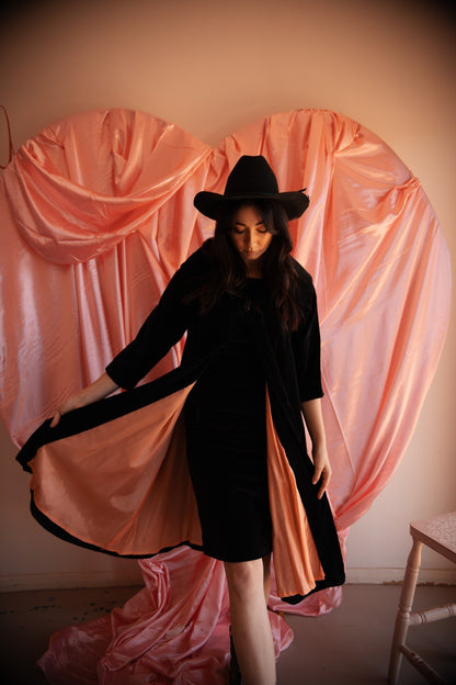 Woman in a black dress and hat standing in front of a pink curtain