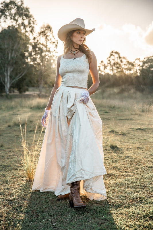 Woman in a white dress and cowboy hat standing in a field with trees in the background