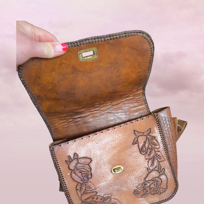 Brown leather bag with floral embossing held by a hand on a pink background