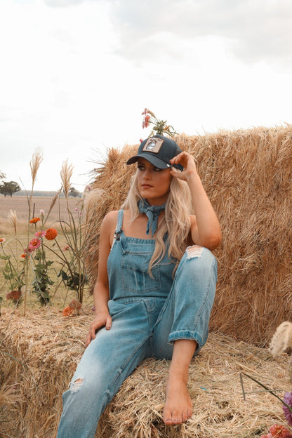Woman in denim overalls and cap sitting on a hay bale with flowers around