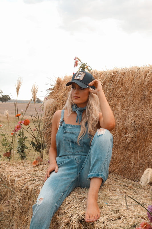 Woman in denim overalls and cap sitting on a hay bale with flowers around