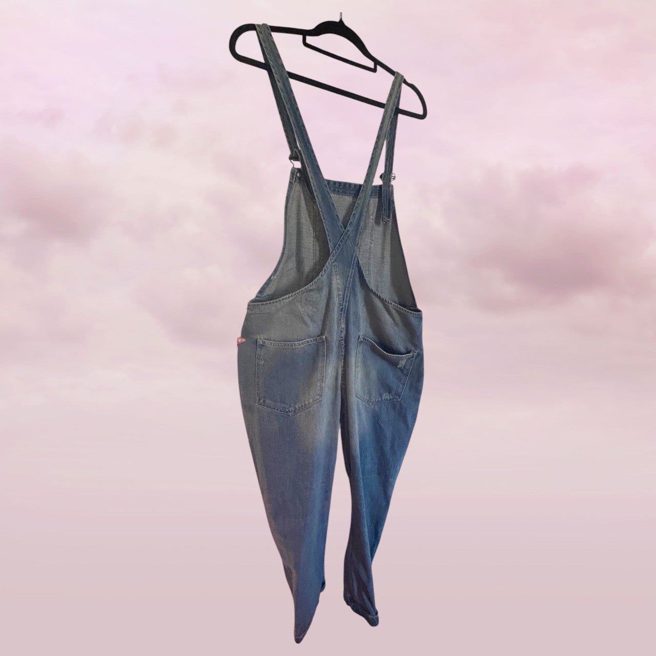 zara distressed Blue denim overalls hanging on a black hanger against a pink background- back view 