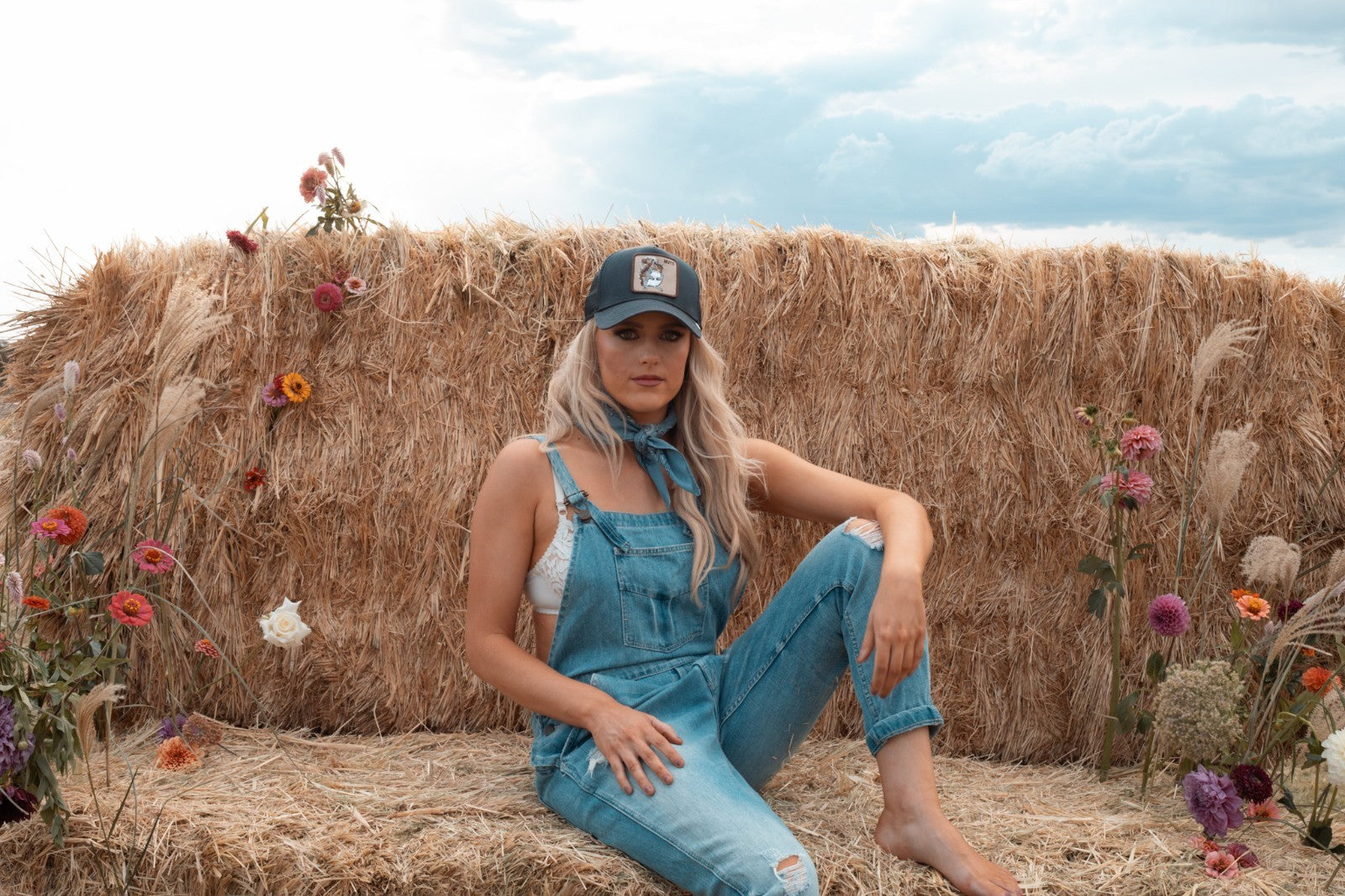 Woman in zara preloved distressed denim overalls sitting on a hay bale with flowers around