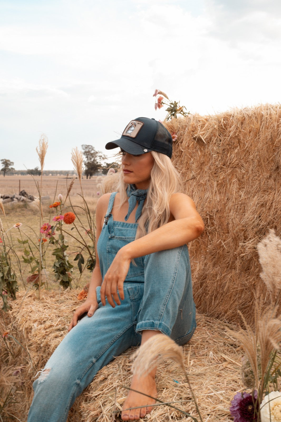 Woman in zara blue distressed denim overalls and cap sitting on a hay bale in a field with flowers.