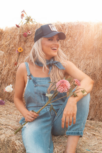 Woman in Zara preloved distressed blue denim overalls and cap sitting on hay with flowers 