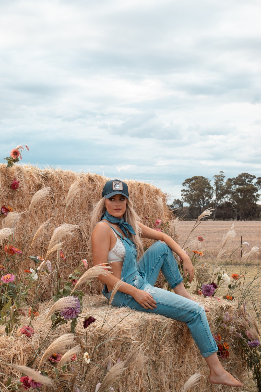 Woman sitting on a hay bale in a field with flowers and a blue cap and zara distressed denim overalls.