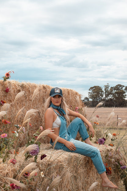 Woman sitting on a hay bale in a field with flowers and a blue cap and zara distressed denim overalls.