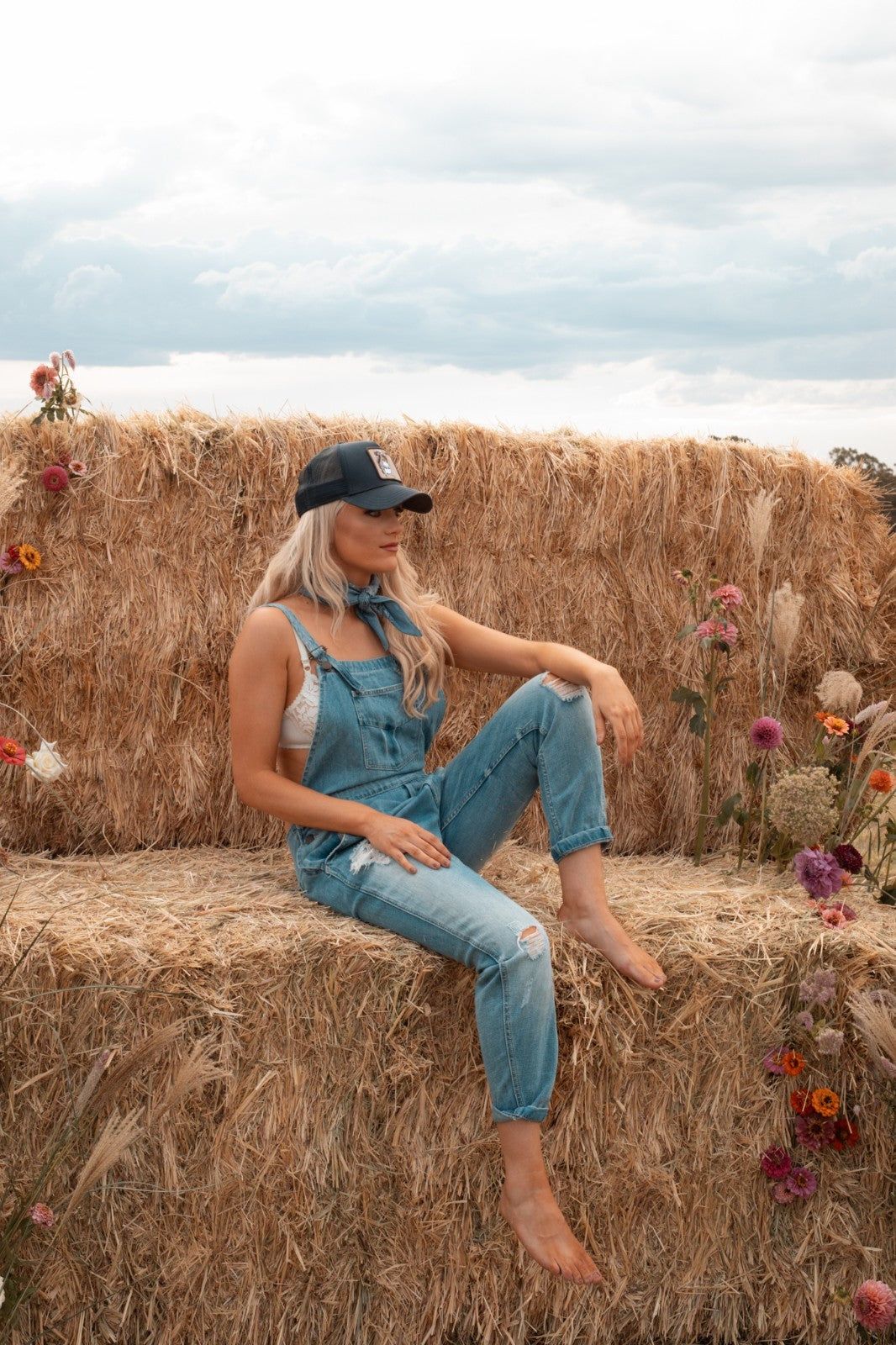 Woman sitting on a hay bale wearing Zara distressed blue zara overalls and trucker capon a hay bale with flowers and a cloudy sky.