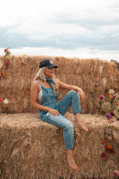 Woman sitting on a hay bale wearing Zara distressed blue zara overalls and trucker capon a hay bale with flowers and a cloudy sky.